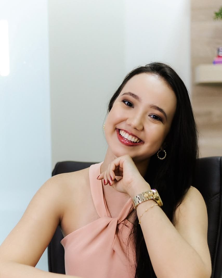 Thamyres B. Cabral, psicóloga, sentada à mesa do consultório, sorrindo com a mão apoiada no queixo.