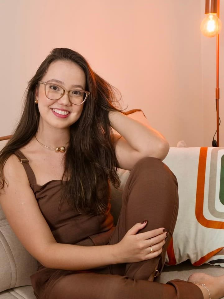 Thamyres B. Cabral de óculos, sentada em um sofá claro, sorrindo em um ambiente acolhedor.